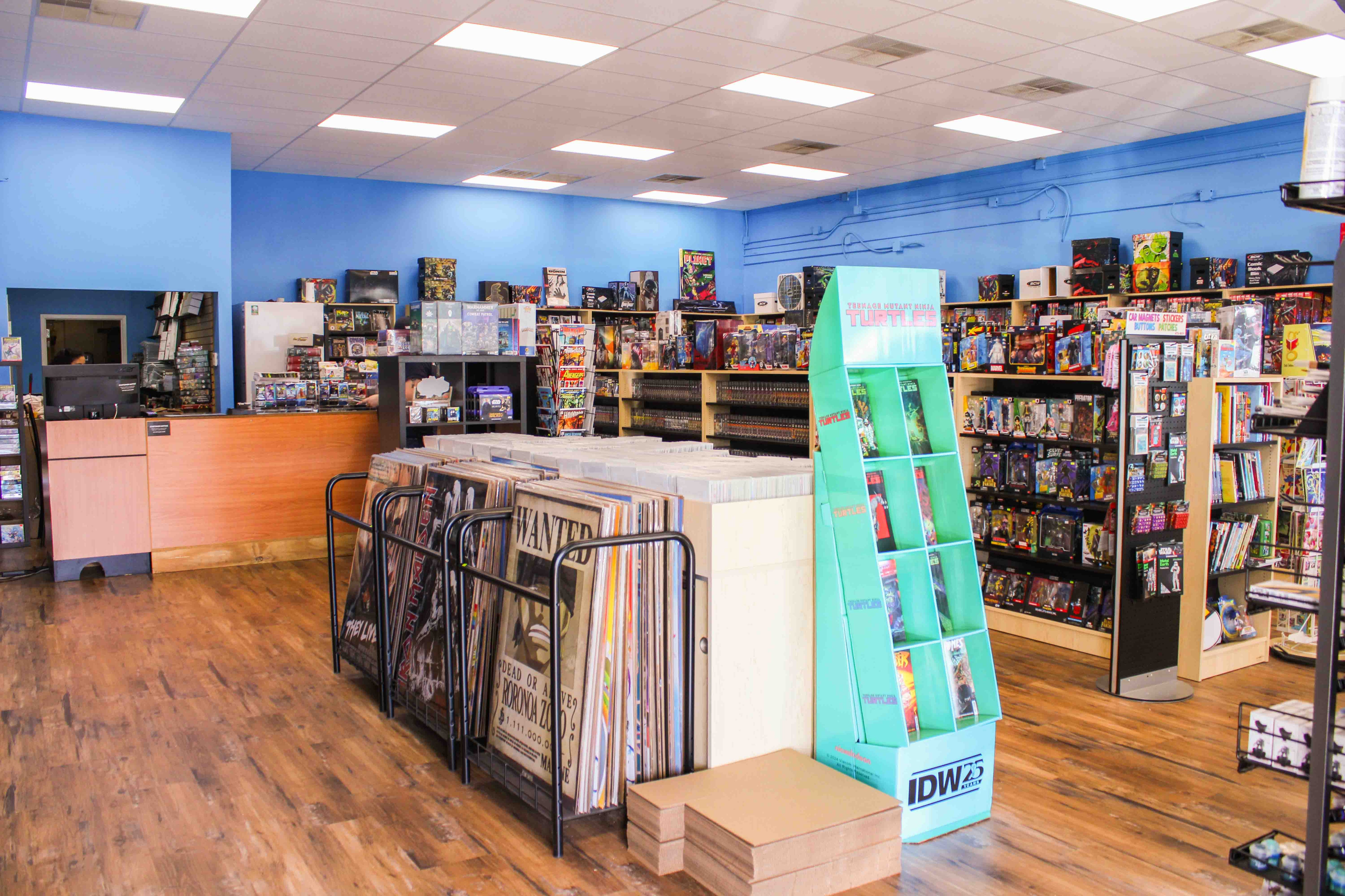 Planet Comics store interior. Shelves of comics and warhammer, poster racks, and back issue comic bins. Image also features front desk with various trading card game booster packs from pokemon, lorcana, and magic the gathering.